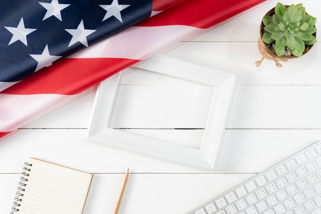 Vista superior del teclado, libro, lápiz, marco de fotos blanco y bandera americana sobre fondo blanco de madera