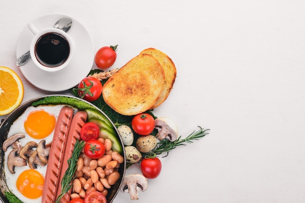 Vista superior de tomates, salchichas, frijoles, champiñones, huevos, tostadas y hierbas para el desayuno inglés. Sobre un fondo de madera blanca.