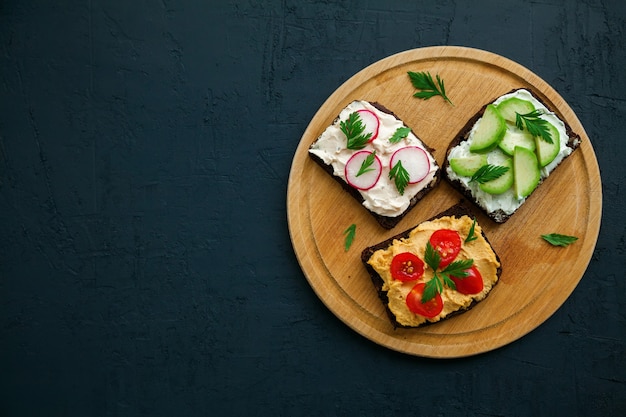 Vista superior de tostadas de pan de centeno vegetariano con requesón, hummus, aguacate, rábano y tomate sobre una tabla de madera, fondo negro, espacio de copia