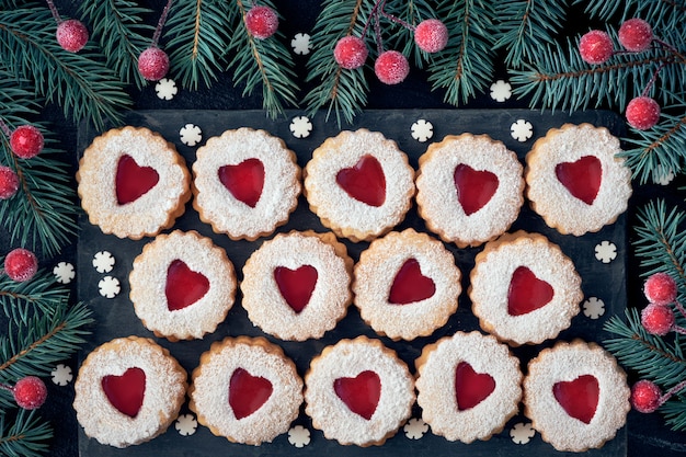 Vista superior de las tradicionales galletas de Navidad Linzer con mermelada roja en la oscuridad decorada con ramas de abeto y bayas