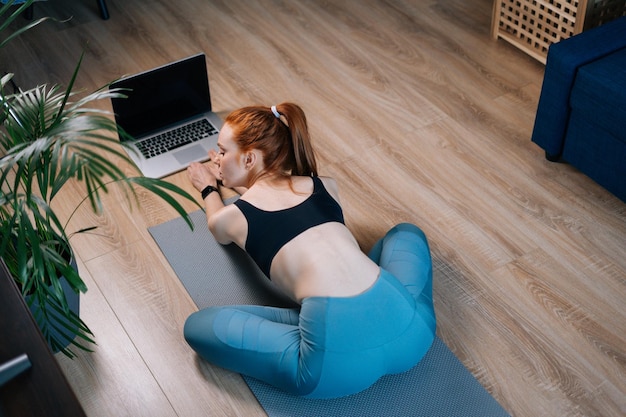 Vista superior trasera de una joven pelirroja haciendo ejercicio, haciendo ejercicio de estiramiento en una alfombra de yoga mientras ve videos de fitness en línea en una laptop. Concepto de entrenamiento deportivo dama pelirroja durante la cuarentena.