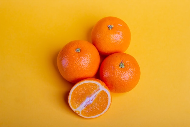 Vista superior tres naranjas frescas sobre fondo amarillo con una naranja partida por la mitad delante, fruta fresca