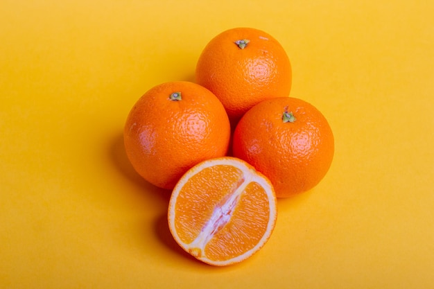Vista superior tres naranjas frescas sobre fondo amarillo con una naranja partida por la mitad delante, fruta fresca
