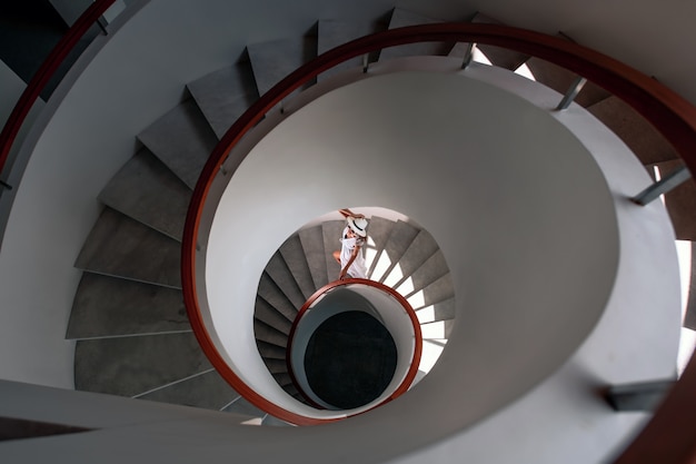Vista superior: uma garota luxuosa de vestido branco e chapéu branco desce uma escada em espiral branca