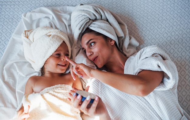 Vista superior. Usando crema. Joven madre con su hija tiene día de la belleza en el interior de la habitación blanca.