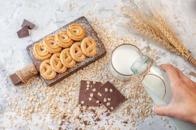 Vista superior de varias galletas sobre una tabla de cortar chocolates espigas de trigo mano vertiendo leche en vidrio avena esparcida sobre fondo gris