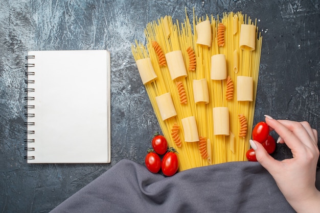 Vista superior de varias pastas rigatoni espaguetis espirales tomate cherry en mano femenina bloc de notas púrpura sobre fondo gris