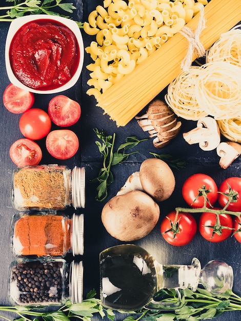 Vista superior de una variedad de pasta cruda cruda junto a un tazón con salsa de tomate, tomates frescos, champiñones, vegetación, diferentes especias y una botella con aceite de girasol