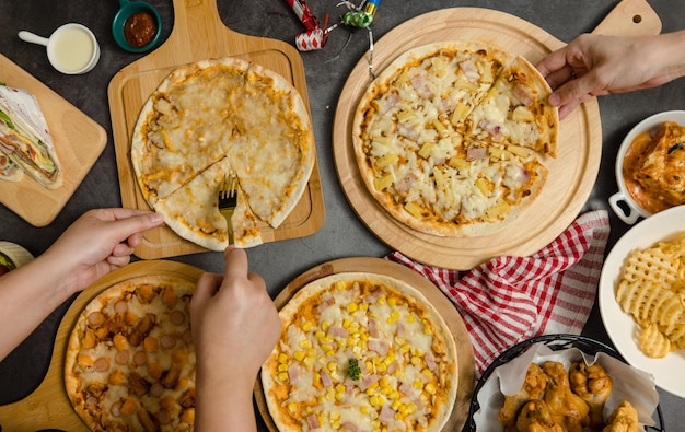 Foto vista superior de varios tipos de pizza deliciosa, sabrosa, jugosa y picante colocada en una tabla de madera en una mesa de fiesta con otra comida rápida mientras que personas irreconocibles recogen la cena juntos