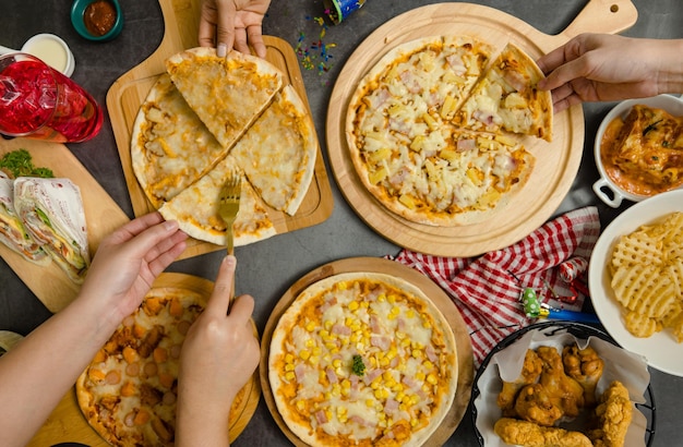 Foto vista superior de varios tipos de pizza deliciosa, sabrosa, jugosa y picante colocada en una tabla de madera en una mesa de fiesta con otra comida rápida mientras que personas irreconocibles recogen la cena juntos