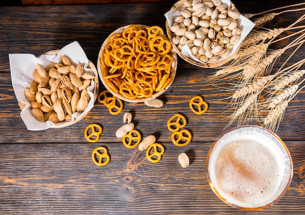 Vista superior del vaso de cerveza con una gran cabeza de espuma cerca de platos con pistachos, pretzels pequeños y cacahuetes en un escritorio de madera oscura. Concepto de alimentos y bebidas