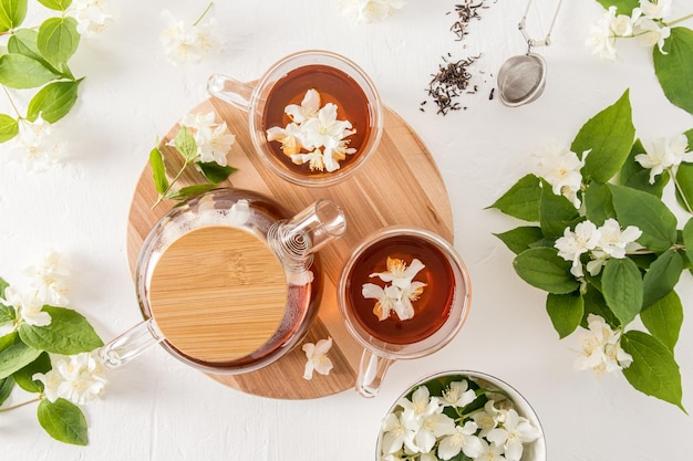 Vista superior de vasos de vidrio y tetera con té de jazmín y flores de jazmín sobre un fondo blanco de tablero de madera redondo