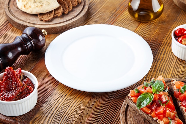 Foto vista superior vazio prato branco para a refeição. prato em branco para servir comida em composição com tomates secos ao sol, bruschetta e utensílios em fundo de madeira. configuração plana