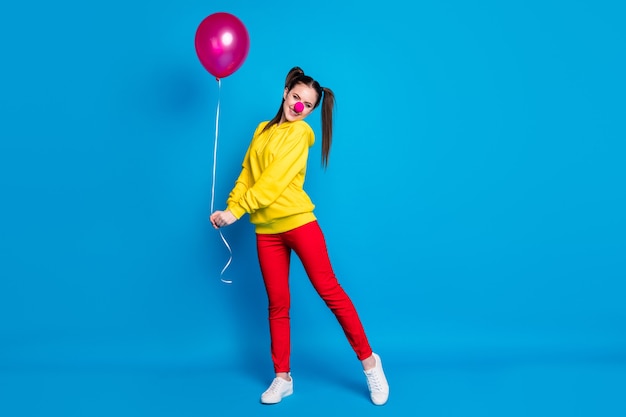 Vista del tamaño del cuerpo de cuerpo entero de agradable atractivo encantado tímido alegre chica alegre payaso de circo sosteniendo en la mano bola de helio divirtiéndose posando aislado sobre fondo de color azul vibrante de brillo vivo brillante