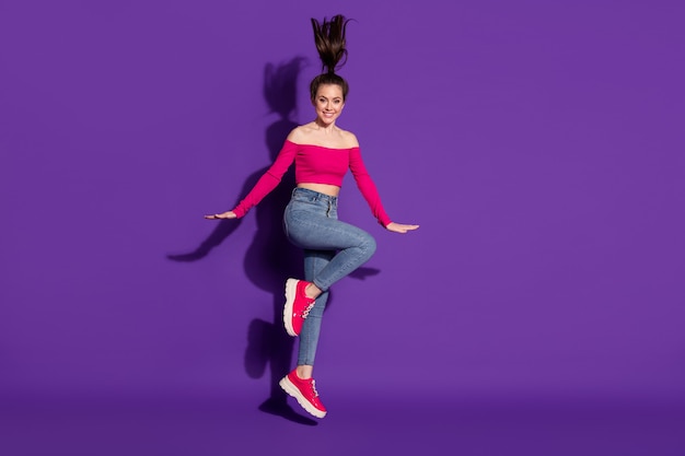 Vista del tamaño del cuerpo de longitud completa de una chica muy alegre que se divierte saltando posando aislada sobre fondo de color violeta brillante