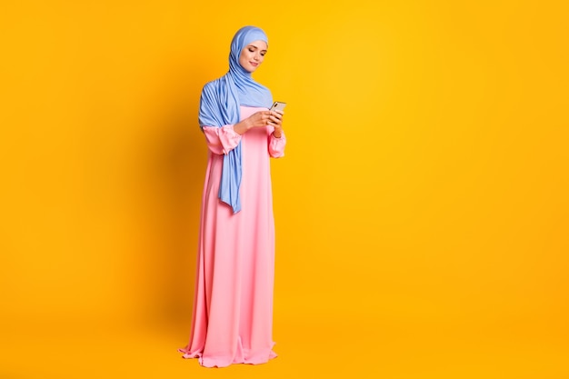 Vista del tamaño del cuerpo de longitud completa de la hermosa muslimah enfocada elegante hermosa que usa el hijab usando la aplicación del dispositivo 5g fondo de color amarillo brillante aislado