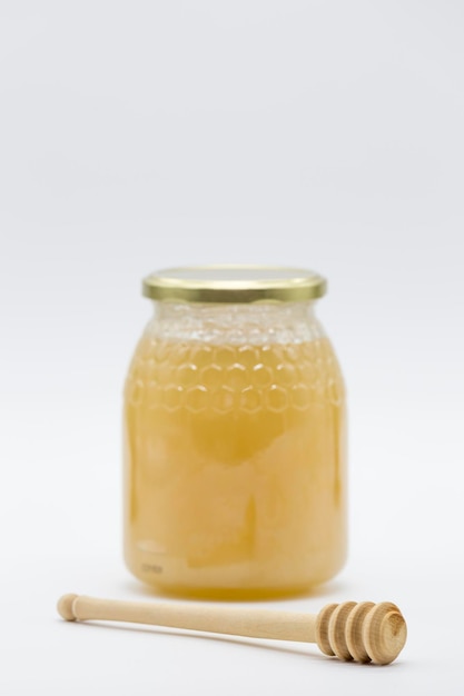 Vista de un tarro de miel junto a una cuchara de miel de madera, todo sobre fondo blanco.