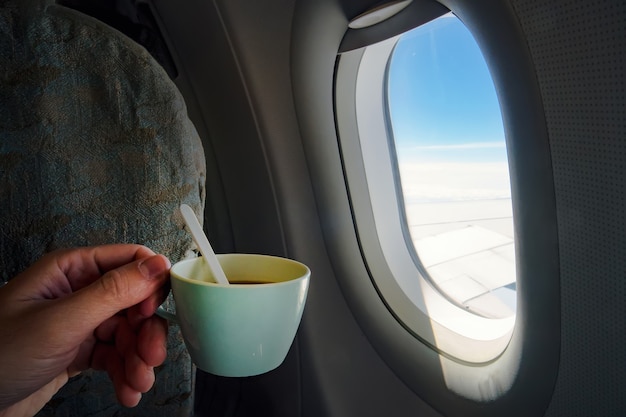 Vista de una taza de café sosteniendo la mano del hombre frente a la ventana de un avión en vuelo en un avión.