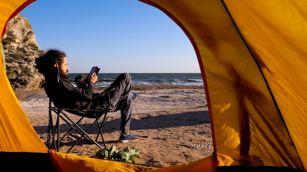 Vista desde la tienda de campaña al hombre barbudo que se sienta en una silla de campamento y lee un lector electrónico en la playa