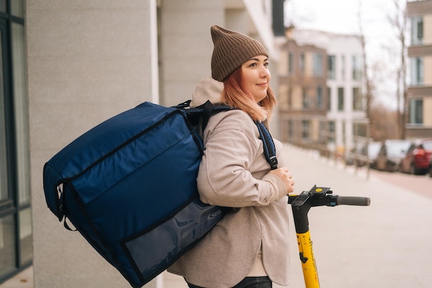 Vista traseira da entrega de comida de correio feminino com grande mochila térmica em pé com scooter elétrico na rua da cidade