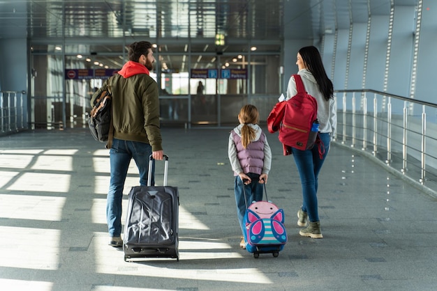 Vista traseira da família de três caminhando ao longo do aeroporto enquanto carregava suas malas enquanto se apressava e tentava não perder seu voo