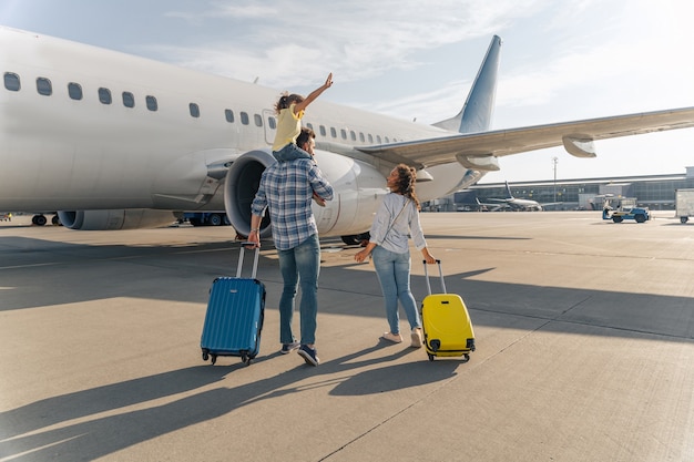 Vista traseira da família feliz em pé perto de um grande avião com duas malas ao ar livre. Conceito de viagem