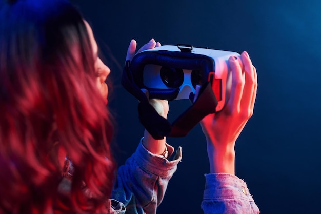 Vista traseira da garota com cabelo encaracolado segurando óculos de realidade virtual em neon vermelho e azul no estúdio