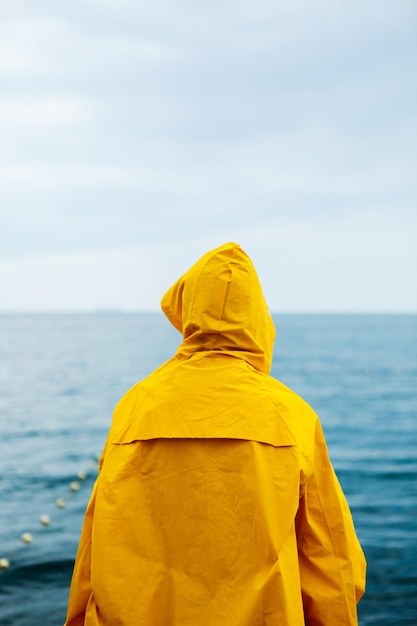 Vista traseira da garota na capa de chuva amarela em pé no cais de pedra à beira-mar com oceano no fundo