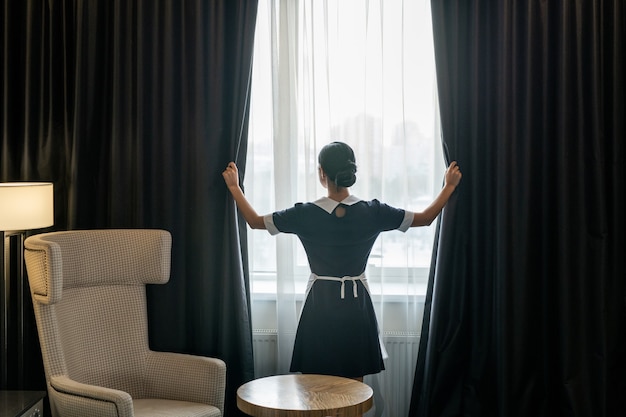 Vista traseira da jovem camareira de uniforme abrindo cortinas escuras penduradas na grande janela do quarto do hotel antes de limpá-la