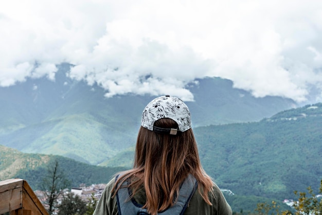 Vista traseira da jovem de boné com camisa xadrez mochila olhando para a vista do céu nublado das montanhas