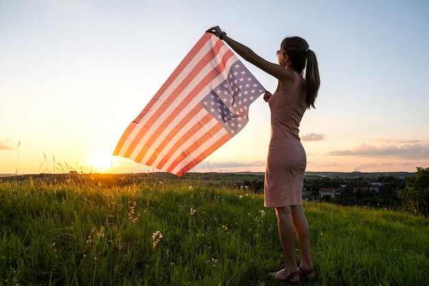 Vista traseira da jovem feliz posando com a bandeira nacional dos EUA ao ar livre ao pôr do sol Mulher positiva comemorando o dia da independência dos Estados Unidos Dia internacional do conceito de democracia