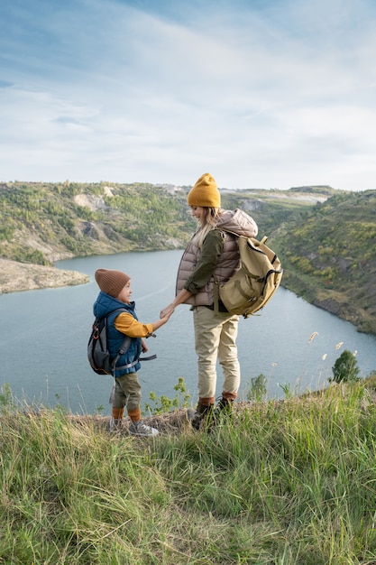 Vista traseira da jovem mãe e filho com mochilas segurando pelas mãos e olhando um para o outro em frente ao lago