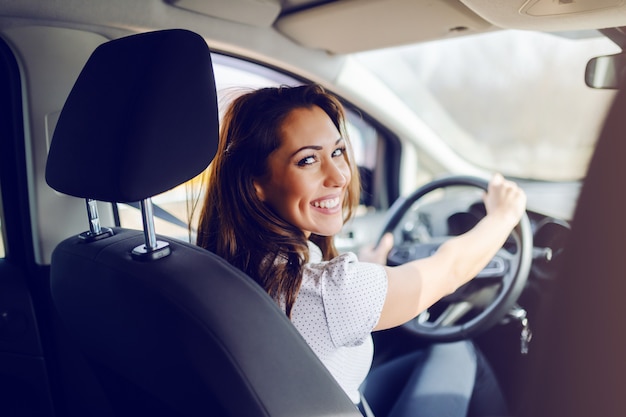 Vista traseira da morena caucasiana sorridente vestido casual inteligente dirigindo seu carro caro enquanto olha para a câmera. As mãos estão no volante.