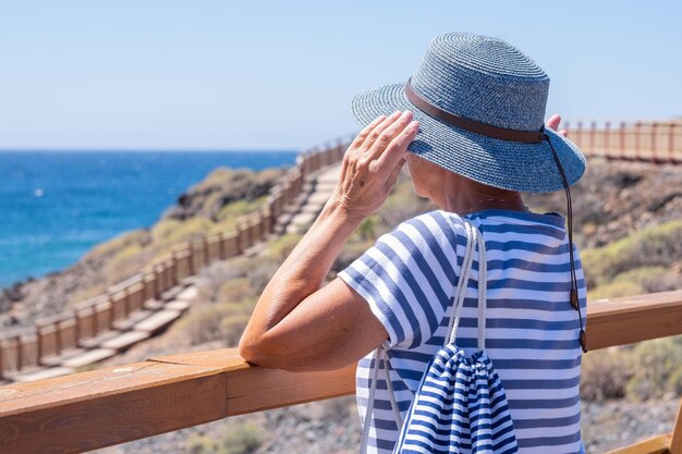 Vista traseira da mulher caucasiana sênior segurando seu chapéu enquanto olha para o horizonte sobre a água aproveitando as férias de verão