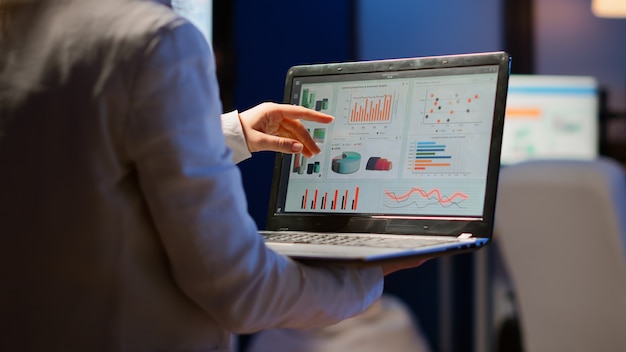 Foto vista traseira da mulher de negócios, analisando as estatísticas financeiras, segurando o laptop em pé no escritório de start-up tarde da noite com excesso de trabalho. funcionário com foco ocupado usando horas extras sem fio de rede de tecnologia moderna