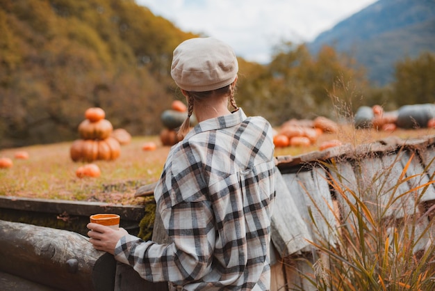 Foto vista traseira da mulher de um agricultor com uma caneca de bebida olhando abóboras em um campo de abóbora contra o fundo das montanhas