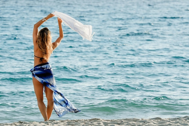 Vista traseira da mulher despreocupada com um xale desfrutando de liberdade de verão na praia