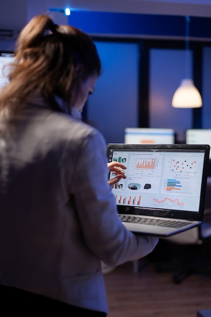 Vista traseira da mulher gerente analisando estatísticas financeiras segurando laptop em pé no escritório de start-up tarde da noite, sobrecarregando