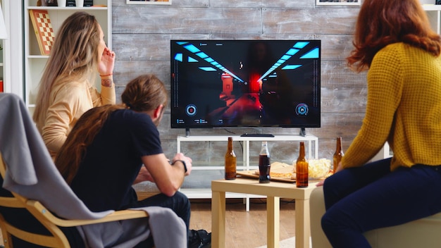 Vista traseira da mulher loira jogando jogos de tiro enquanto seus amigos estão comendo pizza. Jogando jogos com controle sem fio.