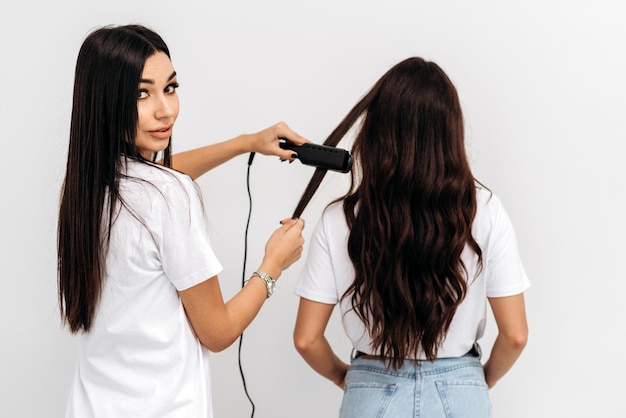 Vista traseira da mulher morena com estilo de cabelo por estilista feminina no salão de cabeleireiro Cabeleireiro profissional olhando para a câmera enquanto passa o cabelo dos clientes