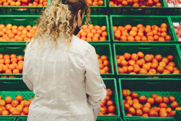 Vista traseira da mulher no mercado comprando frutas ou tomates. Pessoas na atividade de compra de alimentos em uma grande loja. Mulher usando máscara de visão traseira