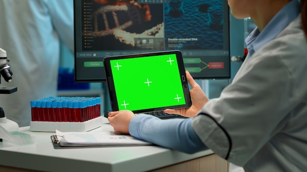 Vista traseira da mulher químico segurando o tablet com maquete verde em laboratório equipado moderno. Equipe de microbiologistas fazendo pesquisa de vacina, escrevendo no dispositivo com chroma key, display isolado.