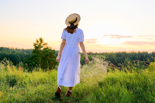 Vista traseira da mulher romântica em um vestido branco com um chapéu de palha apreciando o pôr do sol