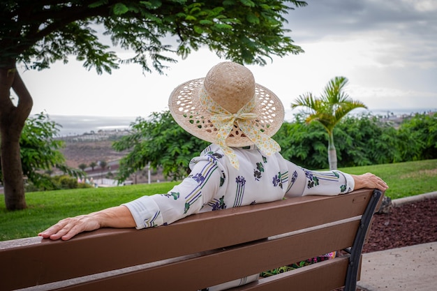 Vista traseira da mulher sênior relaxada com chapéu sentado em um banco no parque público olhando para longe senhora caucasiana aproveitando o tempo livre e aposentadoria