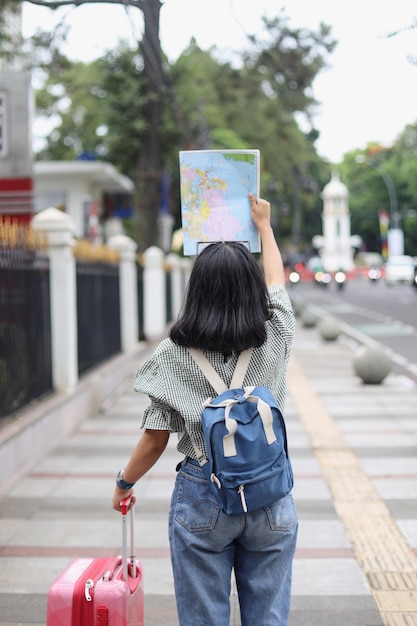 Vista traseira das mãos do viajante feminino segurando o mapa e arrastando a mala e segurando o mapa na calçada