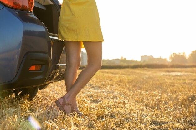 Vista traseira das pernas do motorista jovem com vestido de verão amarelo em pé perto de seu carro.