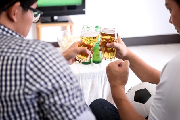 Vista traseira de amigos brindando com garrafas de cerveja enquanto assistem a esportes na televisão em casa