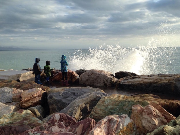 Foto vista traseira de amigos de pé na costa rochosa contra o céu nublado