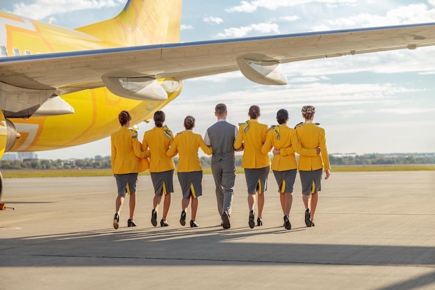 Vista traseira de comissários de bordo de homens e mulheres vestindo uniforme de aviação enquanto caminhavam pelo aeródromo sob a asa do avião