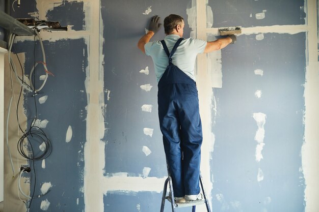 Vista traseira de corpo inteiro de trabalhador da construção civil sênior alisando a parede seca em pé na escada enquanto reforma a casa, copie o espaço